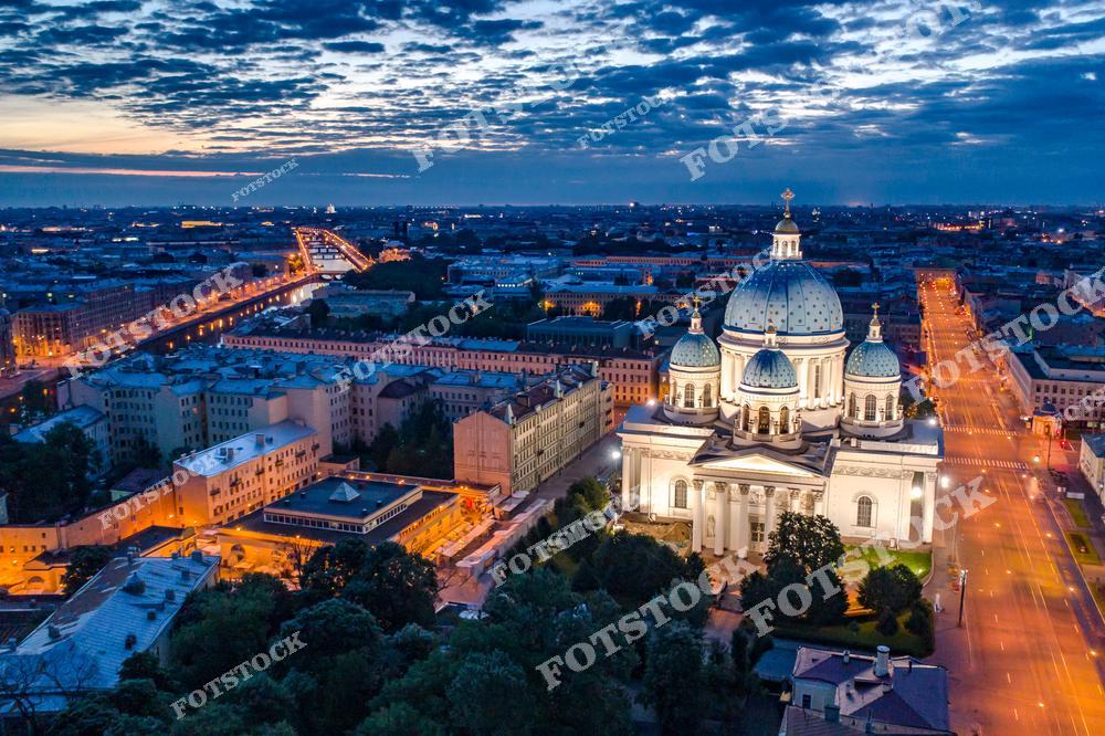 На фотографии запечатлён Троицкий собор в Санкт-Петербурге, подсвеченный огнями ночью. Его белоснежные стены и голубые купола с золотыми звёздами подчёркивают величие храма. На заднем плане видны городской ландшафт и мосты, ведущие через реку Фонтанку. Ночной город создаёт гармоничный ансамбль света и архитектуры.